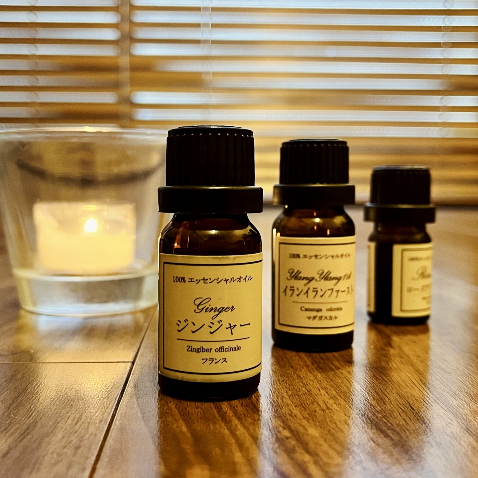 ムーンテッド(MoonTed)の日記より、ジンジャーやイランイランなどの精油の遮光瓶とキャンドル。ブラインドの窓の前