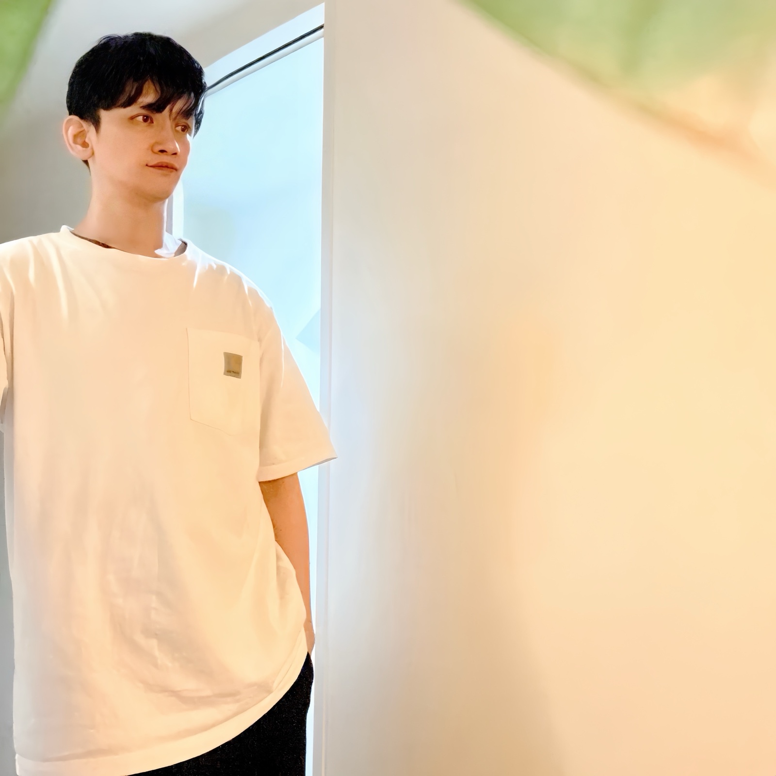 ムーンテッド(MoonTed)の日記より、窓辺に立ち、横を見るムーンテッド(MoonTed)のセラピストAKI。白いTシャツ姿、手前にぼやけた緑の葉
