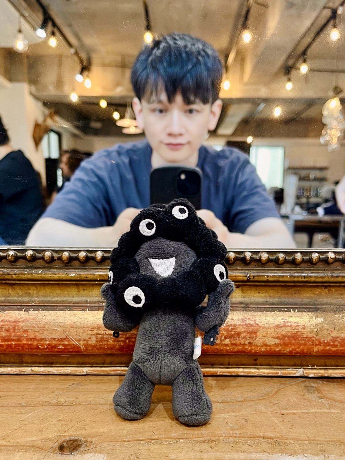 ムーンテッド(MoonTed)の日記より、ヘアサロンの鏡の前に置かれた黒いミャクミャクのぬいぐるみ。鏡にスマートフォンを構えるムーンテッド(MoonTed)のセラピストAKI