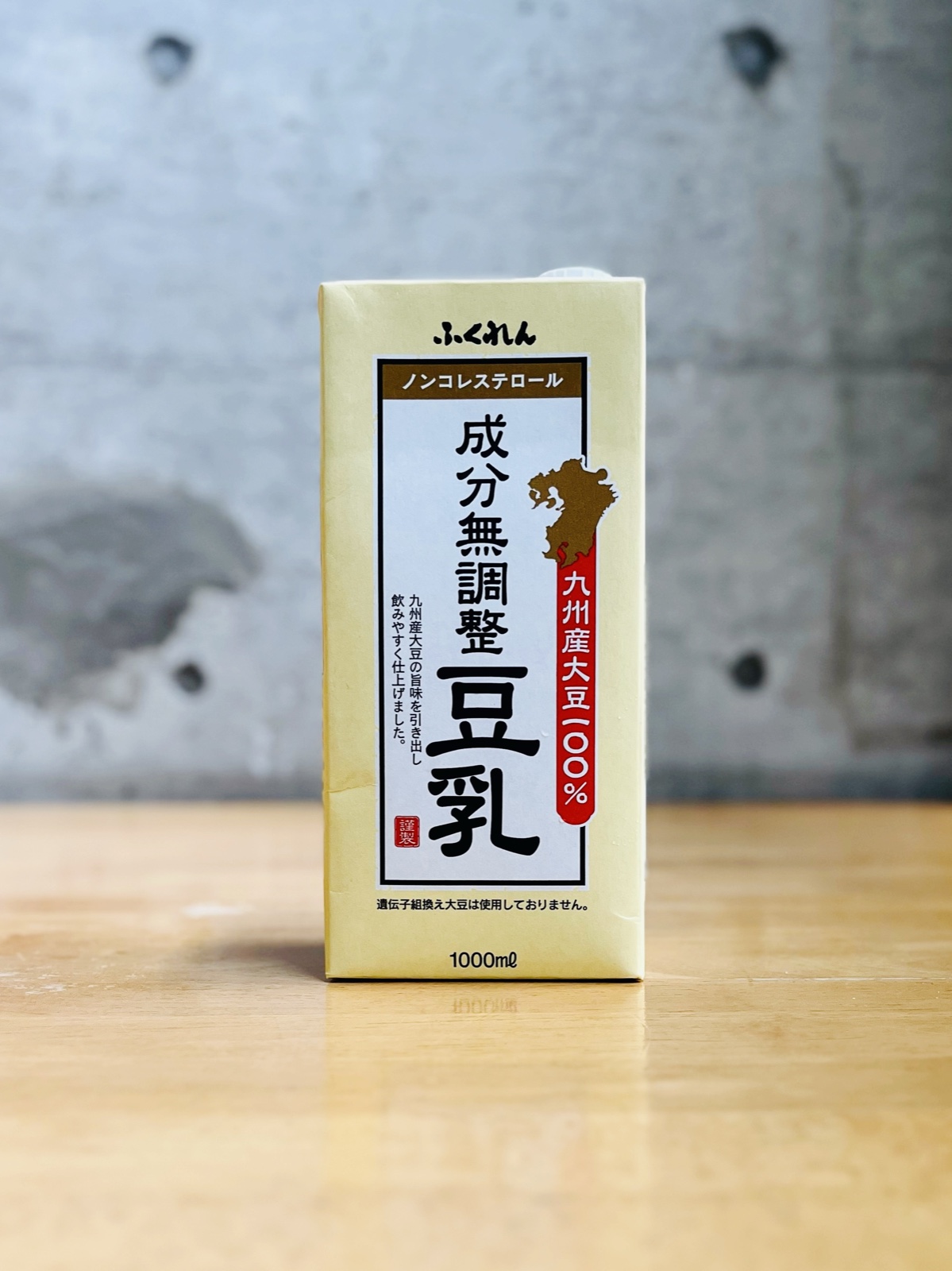 ムーンテッド(MoonTed)の日記より、ふくれんの「九州産大豆 成分無調整豆乳」の紙パック。コンクリート壁の前の木のテーブル
