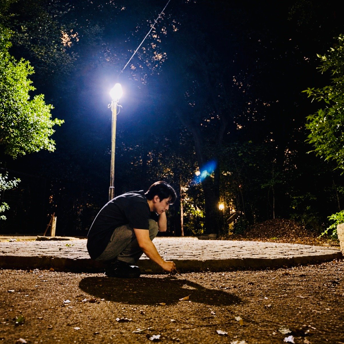 ムーンテッド(MoonTed)の日記より、夜の代々木八幡宮の境内。街灯の下でしゃがみ込み、小枝で地面をつついている男性(AKI)。足元に自分の影が長く伸びている。