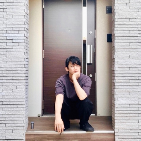 MoonTed Diary: Therapist AKI crouching on the step of a building entrance, chin in hand, in front of a brown door