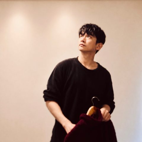 MoonTed Diary: AKI, the therapist, in a black top, holding a wine-red jacket on a hanger and looking up toward the ceiling lights.