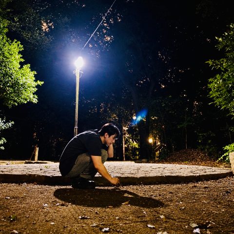 ムーンテッド（MoonTed）の日記より、夜の代々木八幡宮の境内。街灯の下でしゃがみ込み、小枝で地面をつついている男性（AKI）。足元に自分の影が長く伸びている。