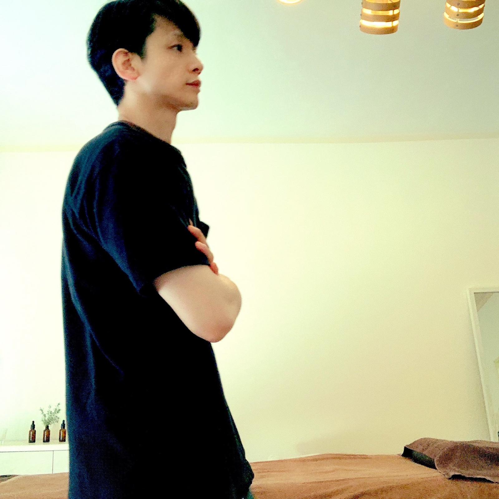 ムーンテッド(MoonTed)の日記より、施術台の横で腕を組んで立つムーンテッド(MoonTed)のセラピストAKI。黒いTシャツ姿の横顔