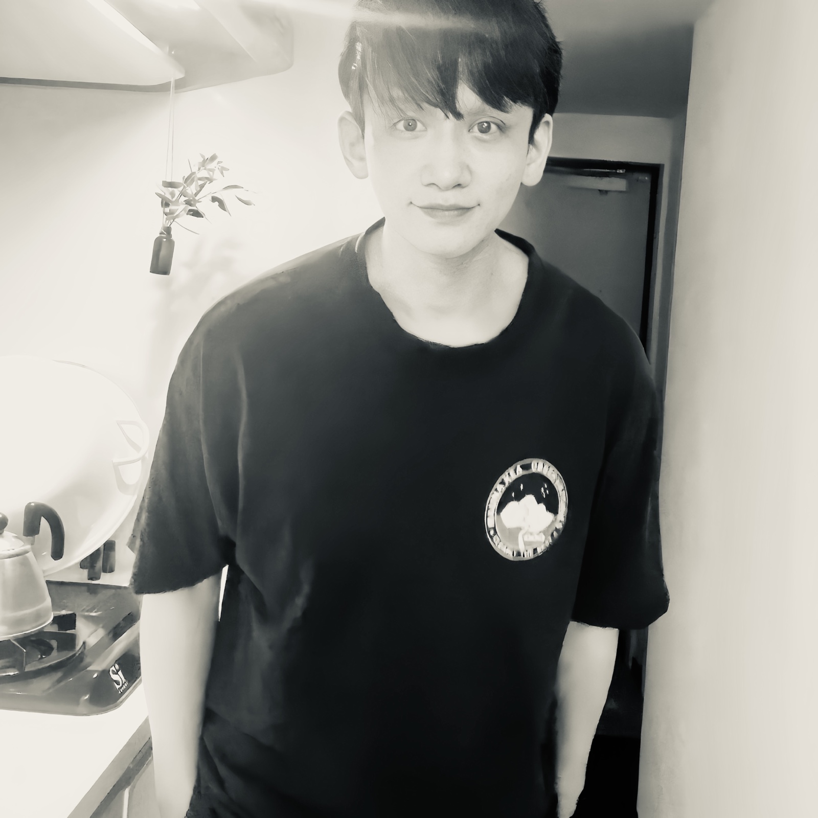 ムーンテッド(MoonTed)の日記より、サロンのキッチンに立ち、少し微笑むムーンテッド(MoonTed)のセラピストAKI。黒いTシャツ姿のモノクロ写真