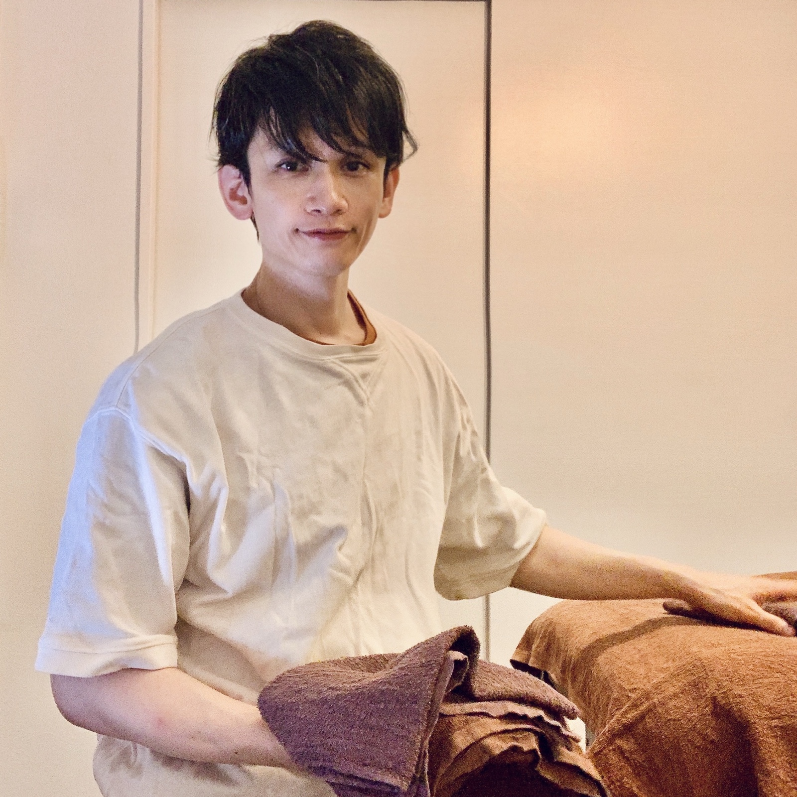ムーンテッド（MoonTed）の日記より、タオルを抱え、施術台に手を置いて微笑むムーンテッド（MoonTed）のセラピストAKI。ベージュのTシャツ姿