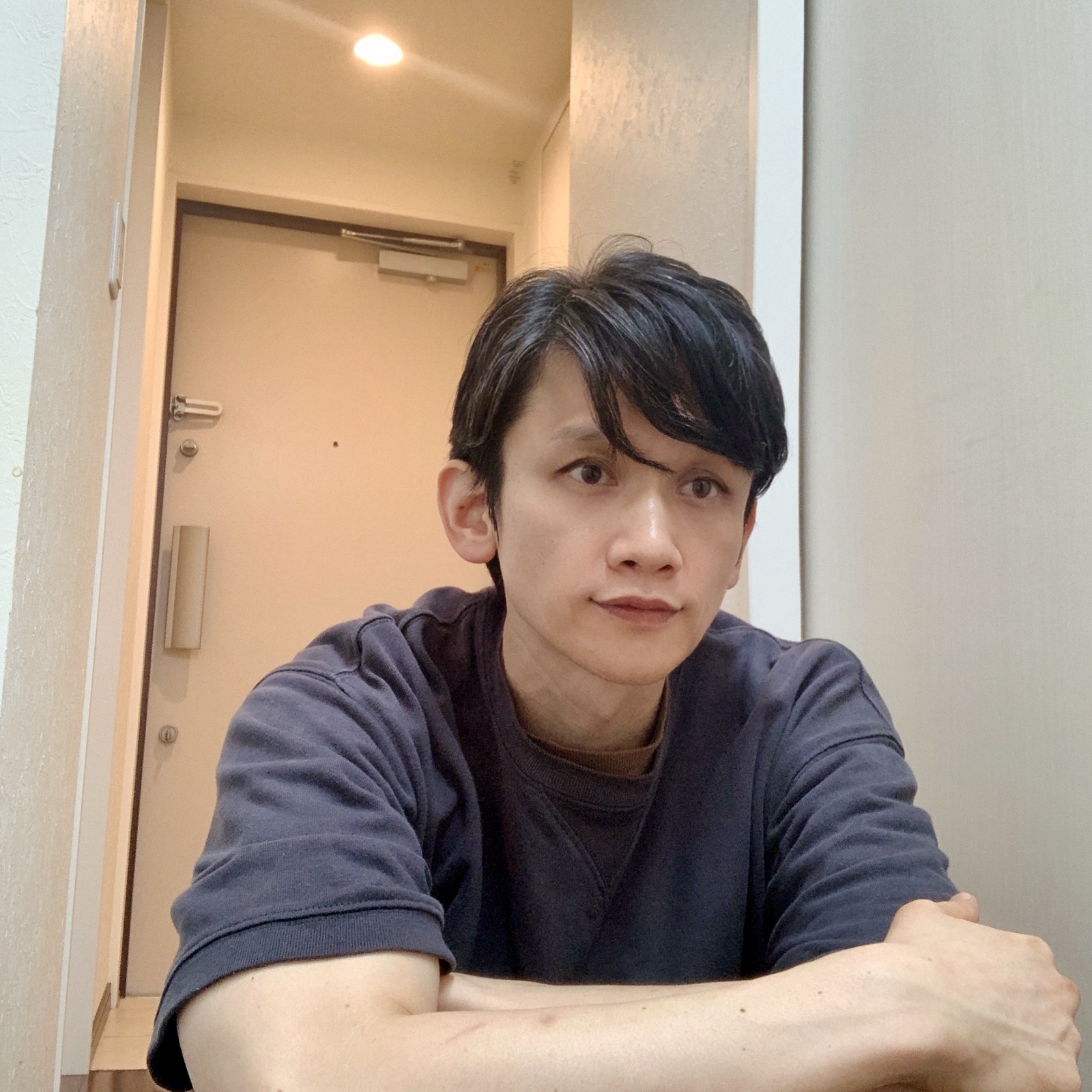 ムーンテッド（MoonTed）の日記より、腕を組んでテーブルにもたれるムーンテッド（MoonTed）のセラピストAKI。紺のTシャツ姿、奥に玄関のドア