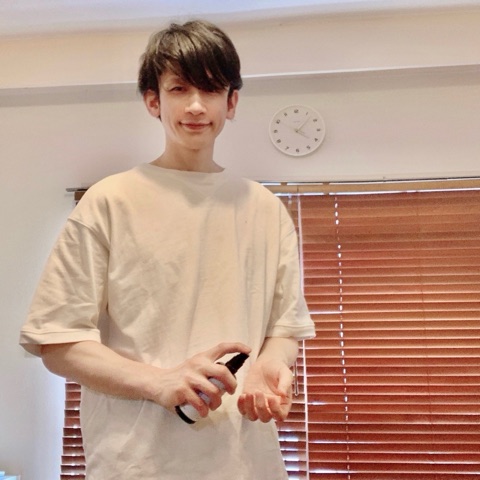 ムーンテッド（MoonTed）の日記より、スプレーボトルを手に立つムーンテッド（MoonTed）のセラピストAKI。ベージュのTシャツ姿、奥に時計とブラインド