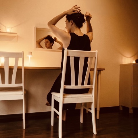 MoonTed Diary: A woman in a black dress on a white wooden chair, tying up her hair in front of a mirror in warm light